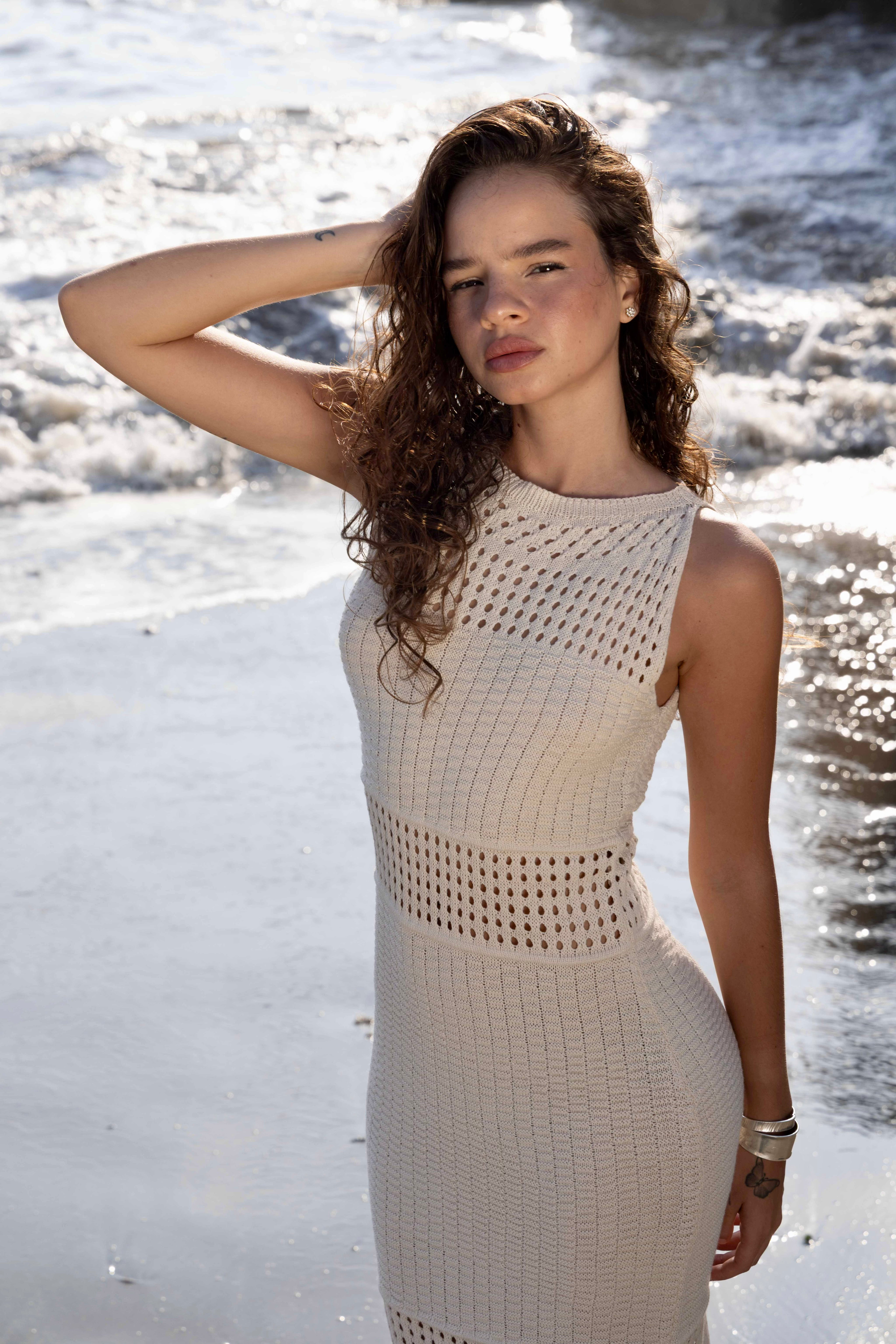 Model wearing Lilian crochet knit maxi dress in cream with high neckline, open-stitch design and fringe hem, walking barefoot on the beach – GYA